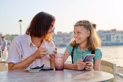 Happy mom and child daughter are resting together. Mother and preteen girl talking laughing in outdoor cafe, seaside city embankment background. Lifestyle, family, friendship, parent and kid concept