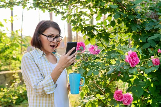 Mature woman resting in the backyard with a cup in her hands using a smartphone for a video call. Lifestyle, technology, middle age people, communication concept