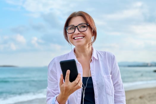 Happy middle aged woman on sea vacation enjoying in headphones with smartphone in her hands. Vacation, lifestyle, technology, leisure, people 40s concept