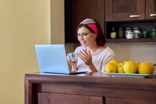 Middle aged woman drinking coffee talking on video call using laptop, kitchen at home. Mature female housewife smiling looking at webcam laptop screen. Technology, lifestyle, home daily life concept