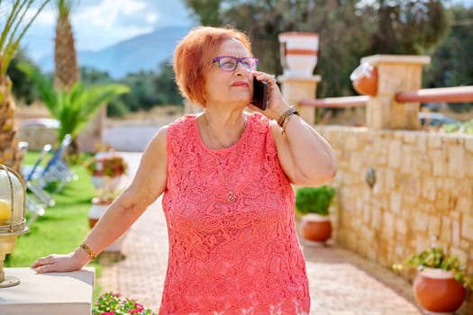 Elderly woman talking on a mobile phone outdoors. 70s retirement female on vacation, summer nature, outside hotel background. Lifestyle, retirees, people concept