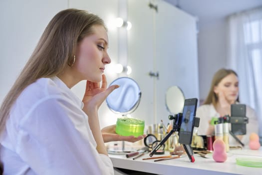 Young woman recording video, makeup beauty blog using smartphone. Female sitting in front of mirror, talking about cosmetics and showing application. Beauty, fashion, care, technology, youth concept