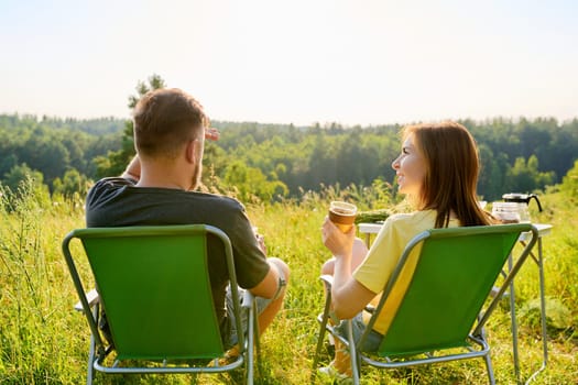 Happy middle aged couple relaxing together outdoor sitting on folding chairs at camping table. Man and woman talking, drinking fresh coffee, enjoying summer nature of wild meadow, back view, weekend