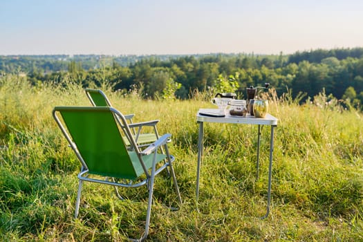 Set of folding furniture for camping, table and chair, cups utensils for brewing fresh coffee, summer nature of wild meadow, sunset on horizon. Summer, holidays, weekends, lifestyle, camping concept