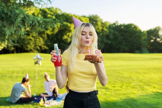 Beautiful teenage girl in festiv hat on birthday with cake and candles. Happy blonde female at outdoor picnic party holding cake with candles 17. Adolescence youth age beauty, holiday birthday concept