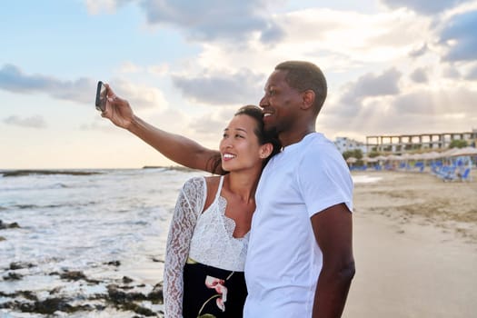 Happy couple in love taking selfie together on smartphone on beach. Vacation tourist trip to seaside resort, summer vacation, multicultural family Asian African American, happiness, date, love concept