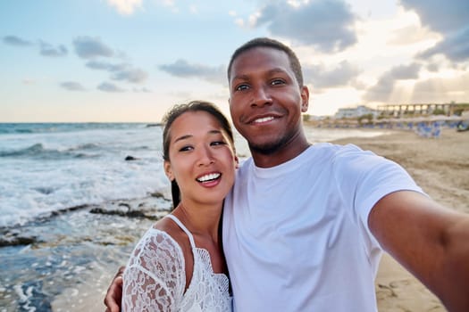 Happy couple in love taking selfie together on smartphone on beach, looking at camera. Vacation tourist trip to seaside resort, summer vacation, multicultural family, happiness, date, love concept