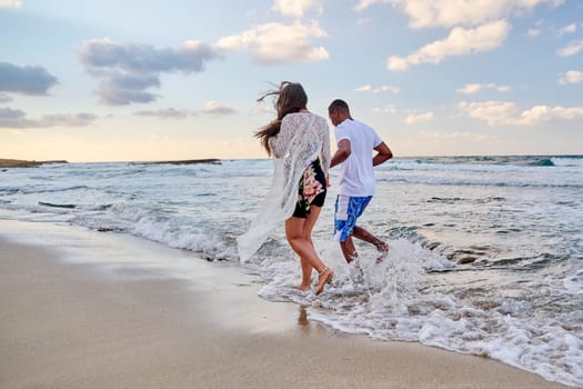 Happy couple running together on the water, on the beach on a summer day, back view. Active young man and woman enjoying sea, nature, vacation, love, relationship, communication concept