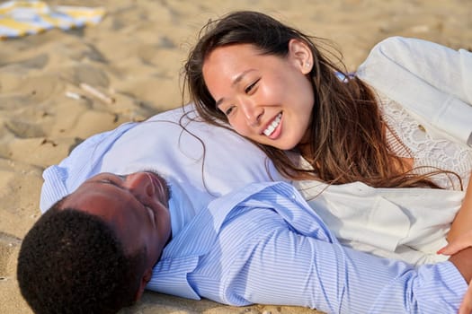 Young happy couple lying on the sand together. Loving multicultural couple, Asian woman and African man, talking enjoying the senses. Love, relationship, family, romance, happiness, people concept