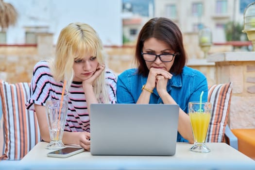 Sad upset serious mom and teenage daughter looking together into a laptop screen. Parent mother and teenage girl sitting outdoor at table with glass of juice watching unpleasant information video.