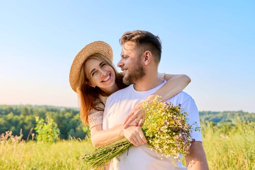 Happy middle-aged couple in love embracing, with bouquet of wildflowers, outdoors on summer nature meadow. Date, anniversary, family, wedding, celebration, relationship, 40s people concept