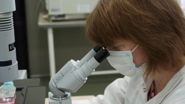 Woman looks through microscope in laboratory. Stock footage. Masked woman looks at biochemical substance through microscope. Analysis of substance under microscope in laboratory.