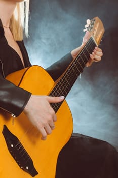 A blonde woman in a black leather jacket plays guitar against a background of smoke