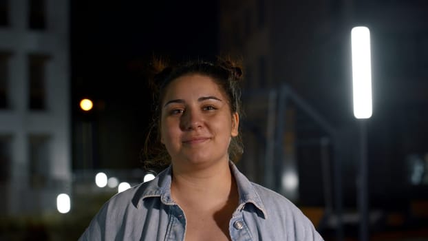 Portrait of attractive caucasian joyful young girl smiling happily to camera outdoors. Media. Dark haired woman with buildings and street lamps on the background winks at camera