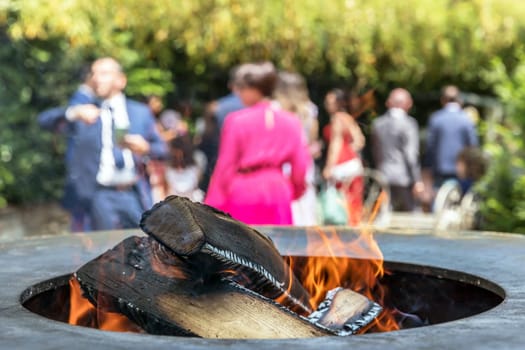 Close up shot of log pieces and fire wood, charcoal and ashes burning in flames in an old vintage brazier. In the blurred background party guests.