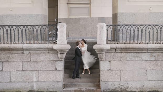 Newlyweds hug on European street. Action. Stylish couple of newlyweds embracing on stone steps of city. Beautiful newlyweds on windy stone embankment of city.