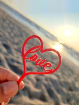 Person holding in fingers hand stick in shape red heart and word Love on background sea and sea waves, blue sky in seashore on sunny summer day close-up. Concept love romance amour St Valentines Day