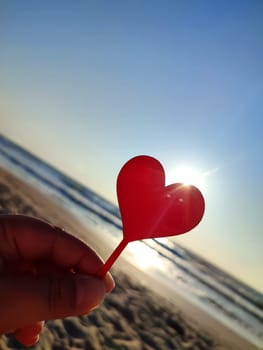 Person holding in fingers hand stick in shape red heart on background sea and sea waves, blue sky in seashore on sunny summer day close-up. Concept love romance amour St Valentines Day