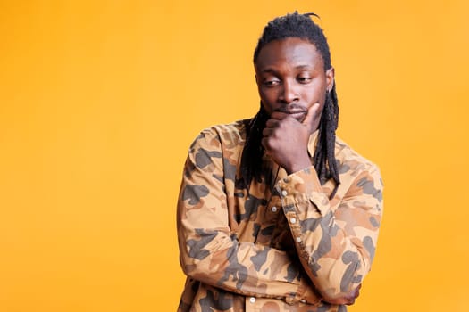 Confused person brainstorming business ideas, thinking at solution to solve difficult problem. Thoughtful african american man having unsure expression, standing in studio over yellow background