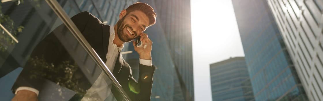 Bottom view of handsome businessman talking by mobile phone standing on background of skyscrapers