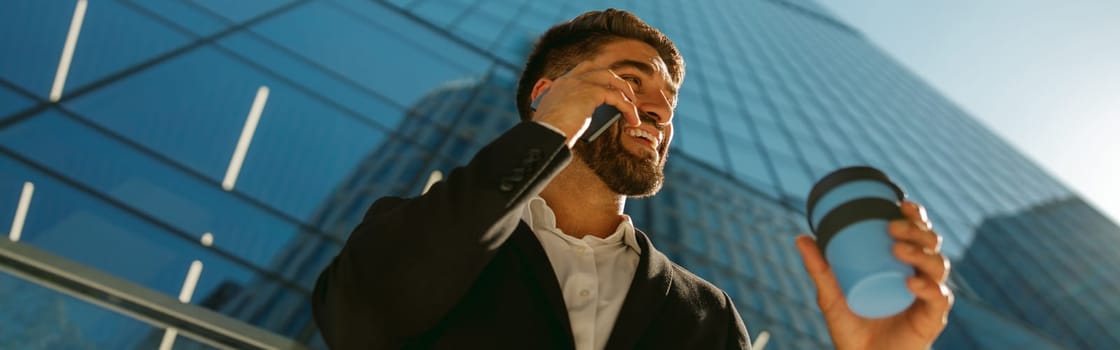 Male manager is talking phone and drinking coffee standing on background of city skyscrapers