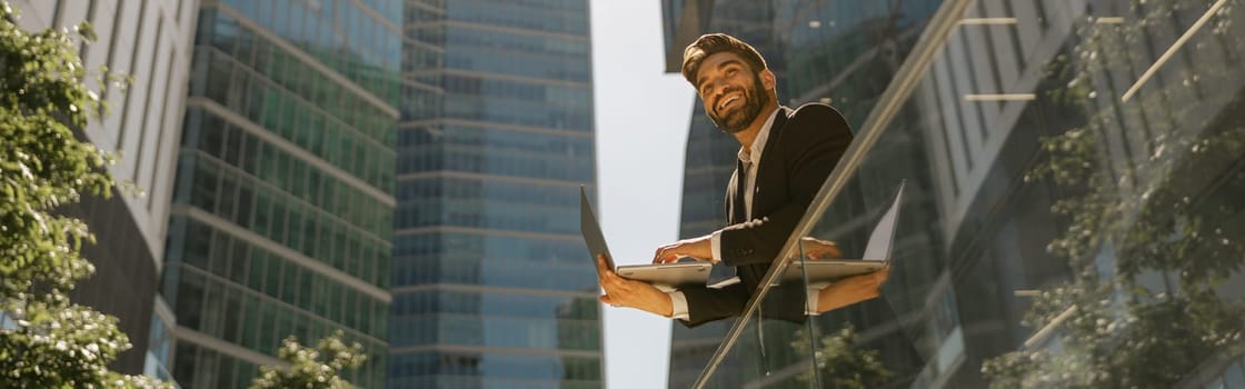 Stylish male manager working on laptop standig on office terrace and looks away. High quality photo