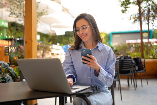 Freelance woman sitting in cafe working with laptop on terrace. Woman wear eyeglasses and smart casual style doing remote work sit on restaurant terrace with nature on background