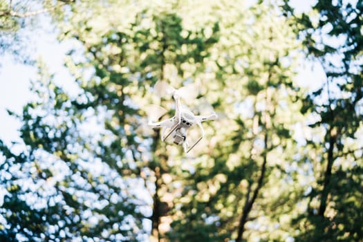 White quadcopter soars in the air against the background of trees in the park. High quality photo