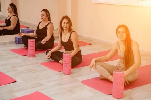 A group of six athletic women doing pilates or yoga on pink mats in front of a window in a beige loft studio interior. Teamwork, good mood and healthy lifestyle concept