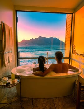 a diverse couple of men and women in the bathtub looking out over the ocean of Cape Town South Africa during vacation. Bath Tub during sunset, multiracial man and woman in bathtub on vacation