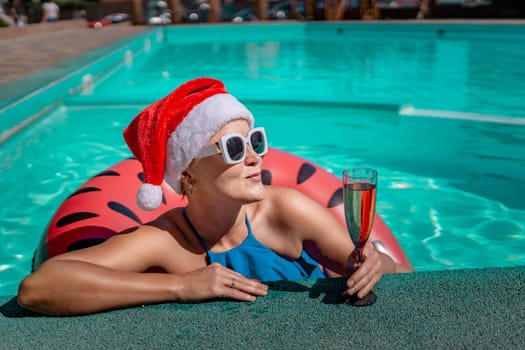 Woman pool Santa hat. A happy woman in a blue bikini, a red and white Santa hat and sunglasses poses near the pool with a glass of champagne standing nearby. Christmas holidays concept