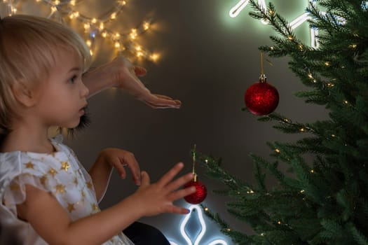 A mother with a 2-year-old daughter decorates the Christmas tree. Mom in a black suit, a girl in a white dress, her daughter hangs a red ball on the Christmas tree. Merry Christmas and New Year concept.