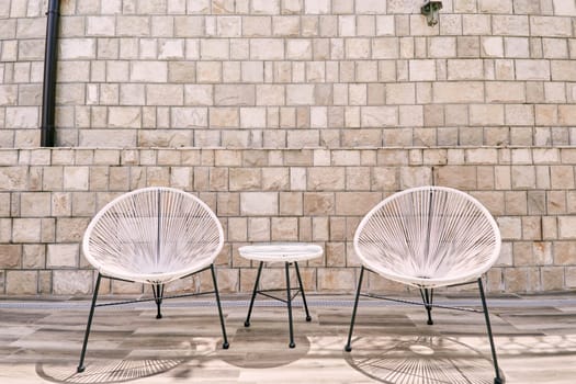 White round polyrattan chairs stand near a table against a stone wall. High quality photo