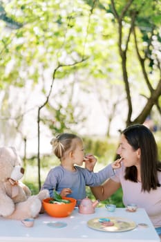 Mom and little girl treat each other with cookies while sitting at a table in the garden. High quality photo