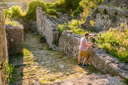 Cameraman in summer outfit with gimbal and cinema camera in his hands making travel video with old europe Balkan nature on the background.