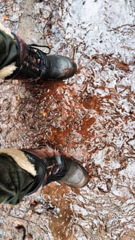 Feet of Hunter or fisherman in big warm boots on winter day on dirty snow. Top view. Fisherman on the river, lake, reservoir on spring day with melting ice. Dangerous fishing in spring or autumn day