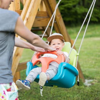 Mother pushing her infant baby boy child on a swing on playground outdoors