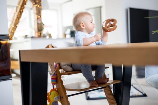 Happy infant sitting at dining table and playing with his toy in traditional scandinavian designer wooden high chair in modern bright atic home. Cute baby playing with toys.