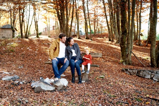 Hugging mom and dad look at a little girl sitting on a bench in the autumn forest. High quality photo