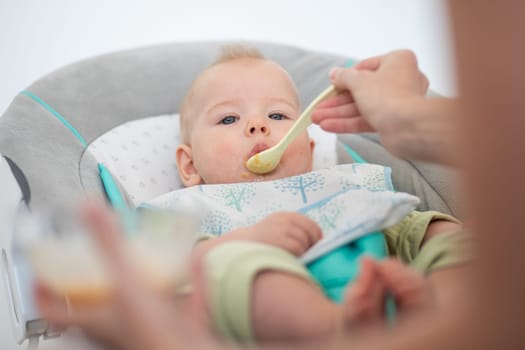 Mother spoon feeding her baby boy child in baby chair with fruit puree. Baby solid food introduction concept