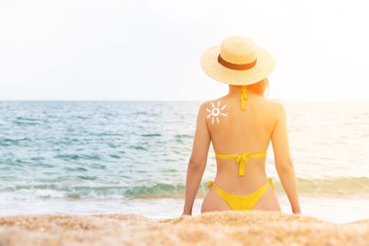 Sun sign on a female body on the shoulder, viewed from the back. Woman sunbathing on the beach with a sun pattern made from sunscreen, sitting near the sea wearing a straw hat.
