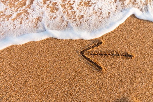 An arrow drawn on the sand pointing to the left next to sea foam.