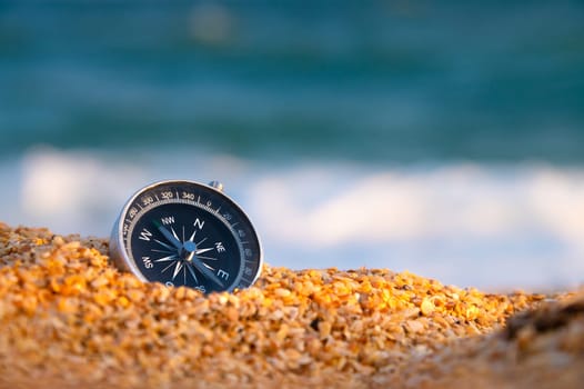 Conceptual photo, close-up of a compass lying in wet sand consisting of small crumbs of shells, against the backdrop of a blue calm sea.