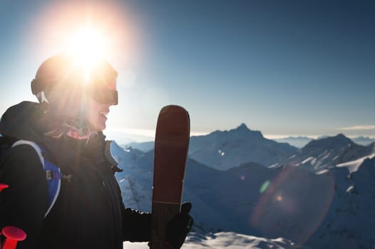 Happy young woman skier against the backdrop of the setting sun and high snow-capped mountains. Portrait of a woman in a protective helmet and ski mask.