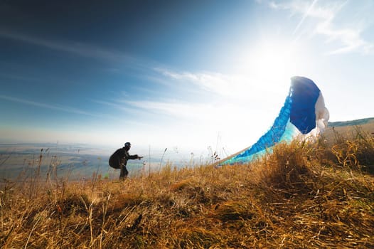 Paragliders take off from a yellow meadow, a man's legs open from the ground, taking off with a parachute upward. Panoramic view of the hills and nature.