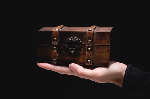The hand of a young Caucasian woman holds a small wooden chest on a black background. Low key.