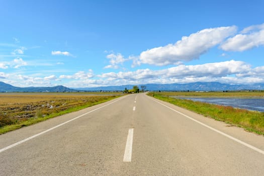 A straight road leading towards mountains under a bright blue sky with clouds, view of lonely road in the ebro delta, tarragona, catalonia, spain,