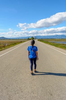 View from behind an individual walking along a deserted road with scenic views on a clear day, back view of latina woman dressed in blue walking down the road in the Ebro Delta natural park, Tarragona, Catalonia, Spain,