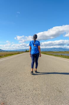 A traveler in casual attire treading along a paved road towards the horizon in a serene setting, back view of latina woman dressed in blue walking down the road in the Ebro Delta natural park, Tarragona, Catalonia, Spain