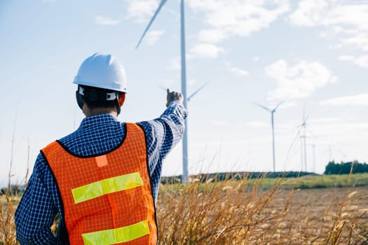 Engineer in hardhat checks windmill farm for clean energy. An Asian technician ensures turbine efficiency. Innovating for global electric security a portrait of expertise and dedication.